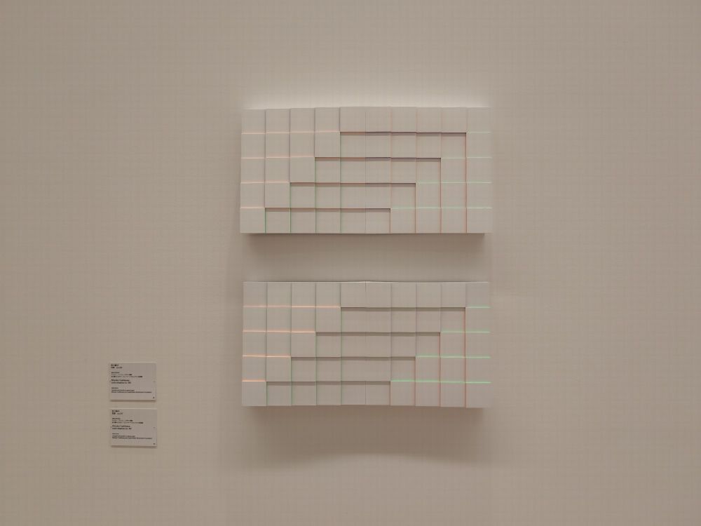 白い壁に展示された二つの幾何学的なアート作品。正方形のパネルが階段状に配置され、細い光のラインが横に走る。左側には作品の説明が書かれたプレートが二枚取り付けられている。