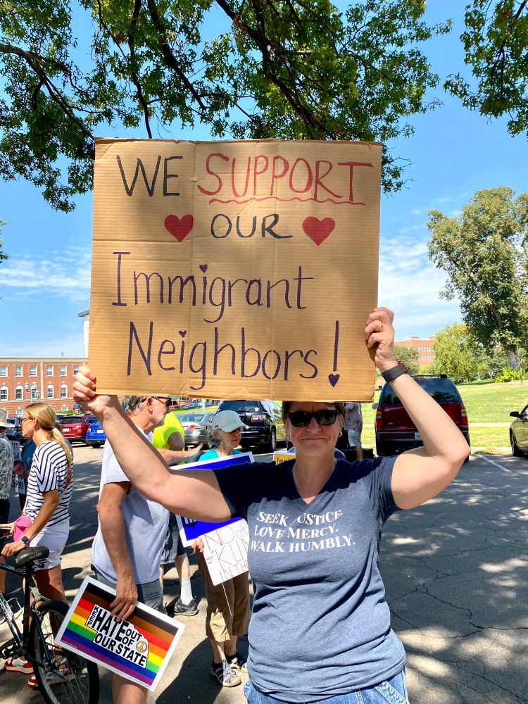 62 year old white woman ( me)  holding a sign that says “We Support Our Immigrant Neighbors!” outside amidst other protestors