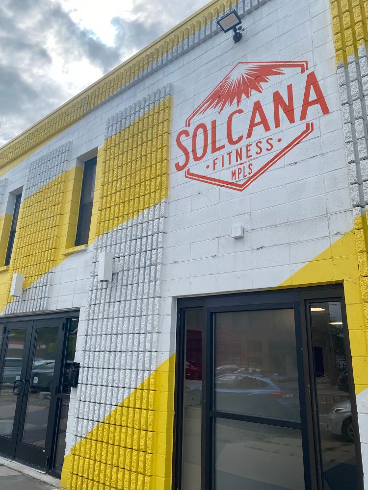 Facade of Solcana Fitness - glass windows and doors are set into white and yellow-striped brick, with an orange logo saying “Solcana Fitness MPLS” on a white background.