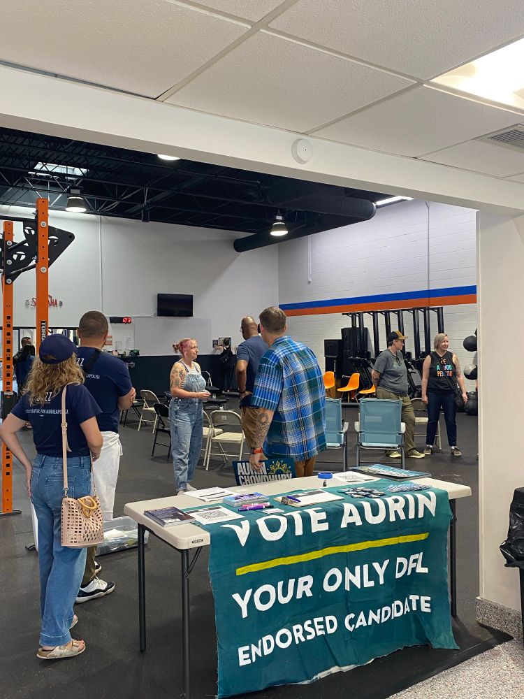 Foreground: a table with a green sign saying “Vote Aurin, your only DFL Endorsed Candidate” and other materials. Multiple people, including Jazz Hampton and DeWayne Davis, are visible in the background talking with each other.