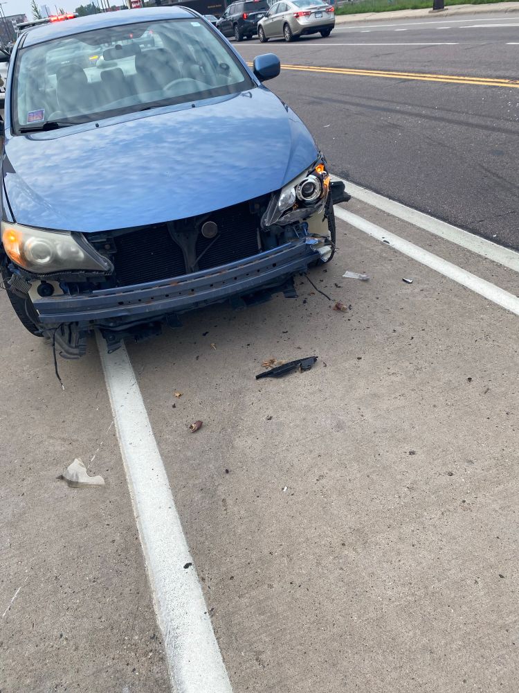 A blue Subaru Impreza is seen with its front bumper torn off, mostly obstructing a bike lane of gray pavement near the black asphalt of car lanes. The driver side rail that holds the bumper is partially destroyed as are the covers for the driver side lights. Various car parts are strewn about the pavement.