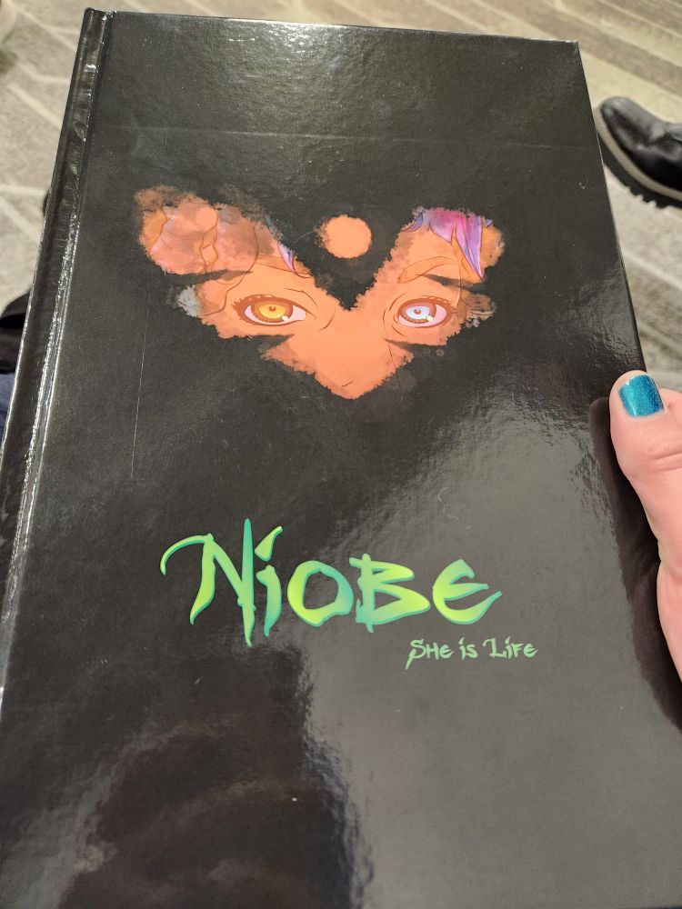 Hand holding a copy of "Niobe: She is Life", a hardcover comic book with a black cover that obscures a woman's face except for a small V-like pattern in the center. Her eyes are yellow and blue, she is dark-skinned and you can see a hint of thick curls and purple leaves.