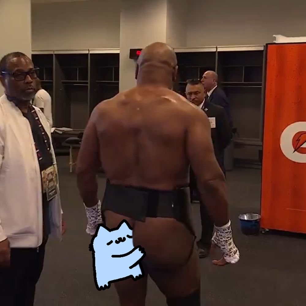 A muscular figure, identified as Mike Tyson, stands in a sports setting, facing away from the camera. He is wearing boxing attire. To his left, another person in a white jacket is partially visible. The background features a red sign with a partial logo, and a cartoon cat character is superimposed at the bottom of the image."