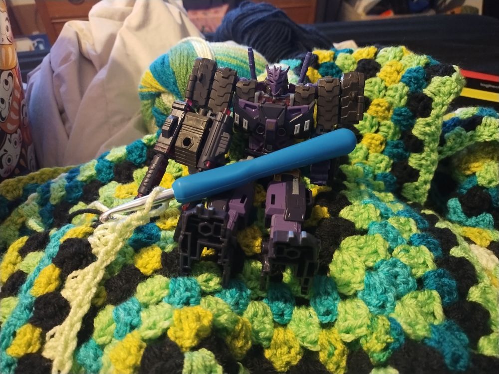 a photograph of a toy of the transformer character tarn sitting on top of a bright green crochet project. there is a blue crochet hook on the figures lap