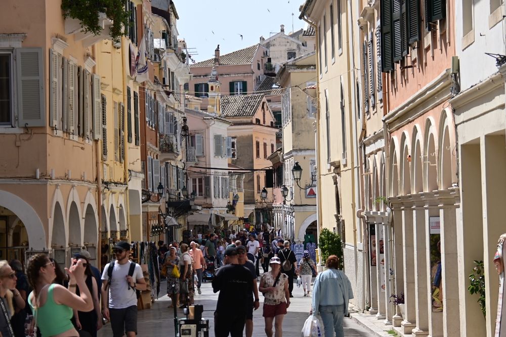 Busy street in Corfu Town. Narrow street full of tourists  tall shop buildings on either side with arched entrances  pastel colours