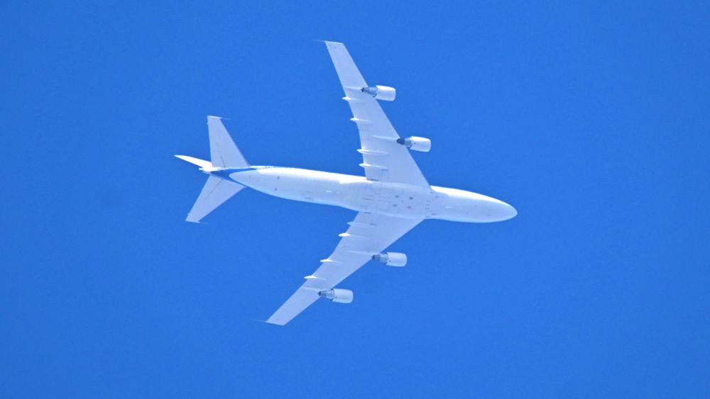 Underside of low flying large white Boeing passenger jet with four engines framed against clear blue sky. Tail has blue markings on underneath. Flight shows up as Trunk72 Omaha to Lakenheath