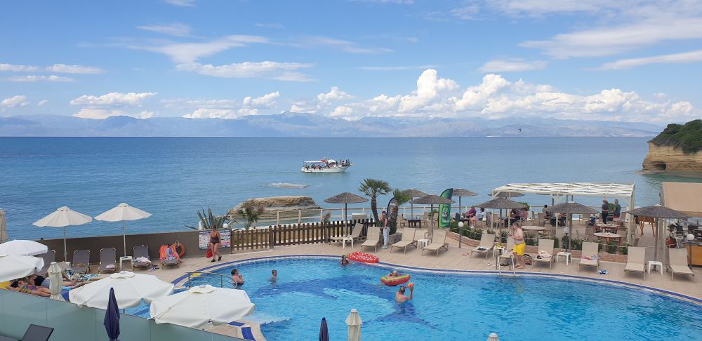 Sunny view in Corfu. Looking out across swimming pool with sun loungers. Ionian sea behind with mountains of Albania on horizon. Blue skies with white clouds over mountains. Small pleasure boat close to shore. Rocky outcrop on right.