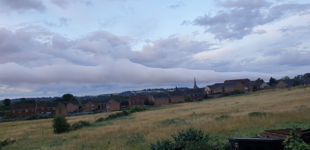 View across a field in late evening with houses in distance, above them is what appears to be a roll cloud stretching width of eastern horizon.