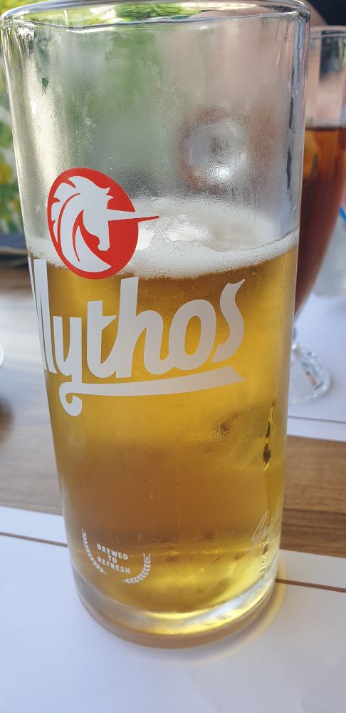 Glass of beer with Mythos logo. Glass is half full of golden lager an misted with condensation.