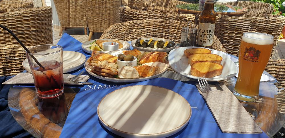 Greek meso meal spread on a table in dappled shade  soft drink on left, local Corfu beer on right, wicker chairs in background, empty plate in foreground on blue table mat.