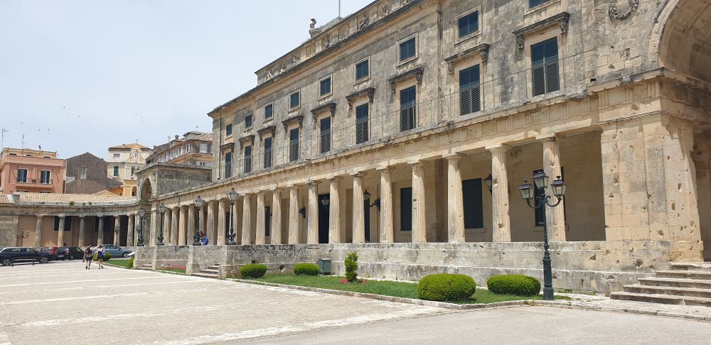 Buildings of Corfu fort. Yellow stone building, frontage of lower floor has columns, two further floors on top with rectangular windows. Courtyard in front with some low hedges.