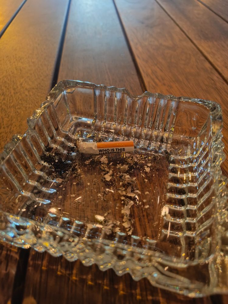 Close-up image of a clear glass ashtray on a wooden table, containing ashes and a cigarette butt with the text 'WHO IS THIS' printed on the cigarette butt. The angle of the photo hides the actual Australian government warning message 'WHO IS THIS HARMING?'