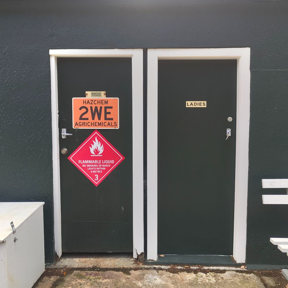 Two side-by-side doors on a dark green building; the left door has hazardous material signs indicating 'Hazchem 2WE Agrichemicals' and 'Flammable Liquid – No Smoking or Naked Lights within 3 Metres,' while the right door is labeled 'Ladies.'