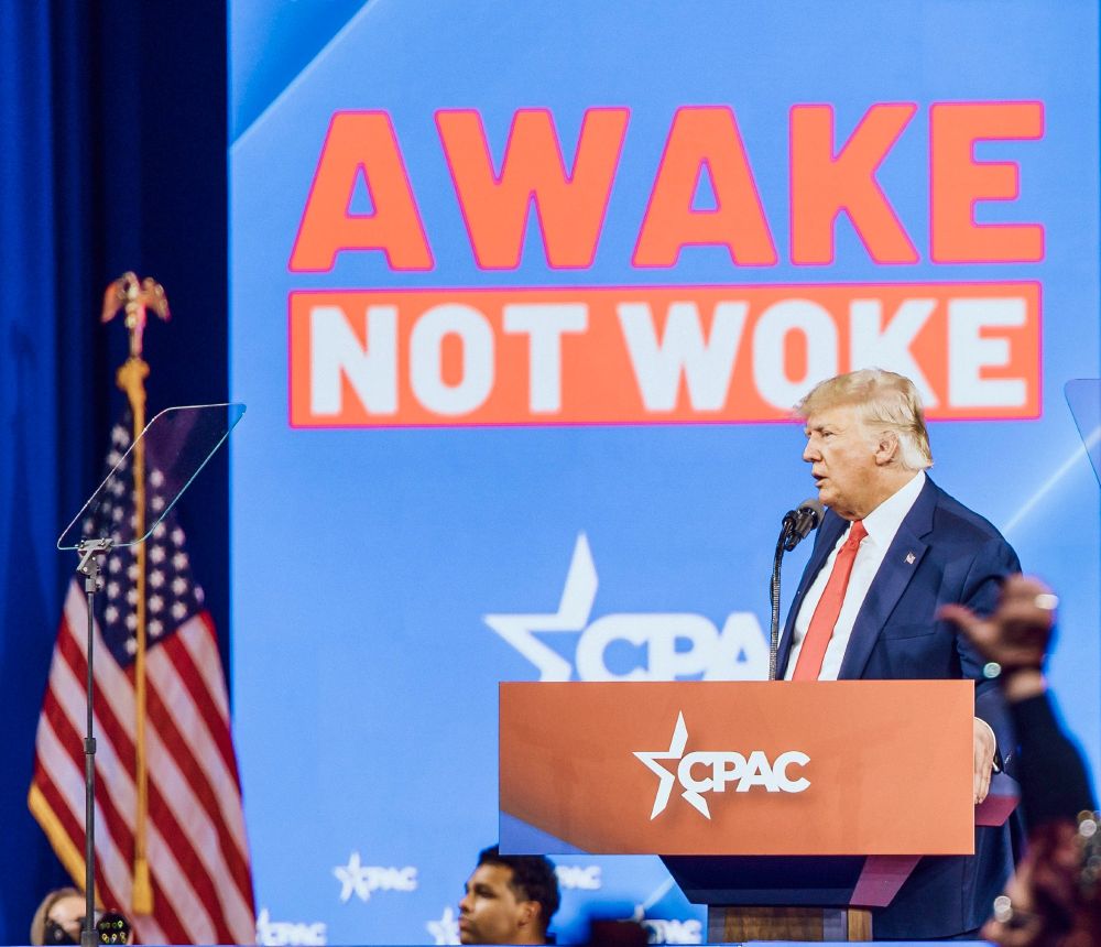 "Donald Trump Speech Awake Not Woke Slogan" - Donal Trump est à côté du drapeau américain, derrière un pupitre estampillé "CPAC", avec derrière lui, sur le mur, écrit en gros "AWAKE" puis "NOT WOKE"