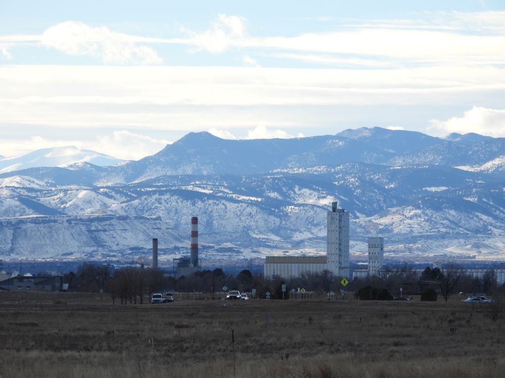An industrial facility, far away, red and white smokestacks, with extreme zoom, making the mountains appear to be twice as tall as the buildings, looming impassibly overhead, snowy and forbidding