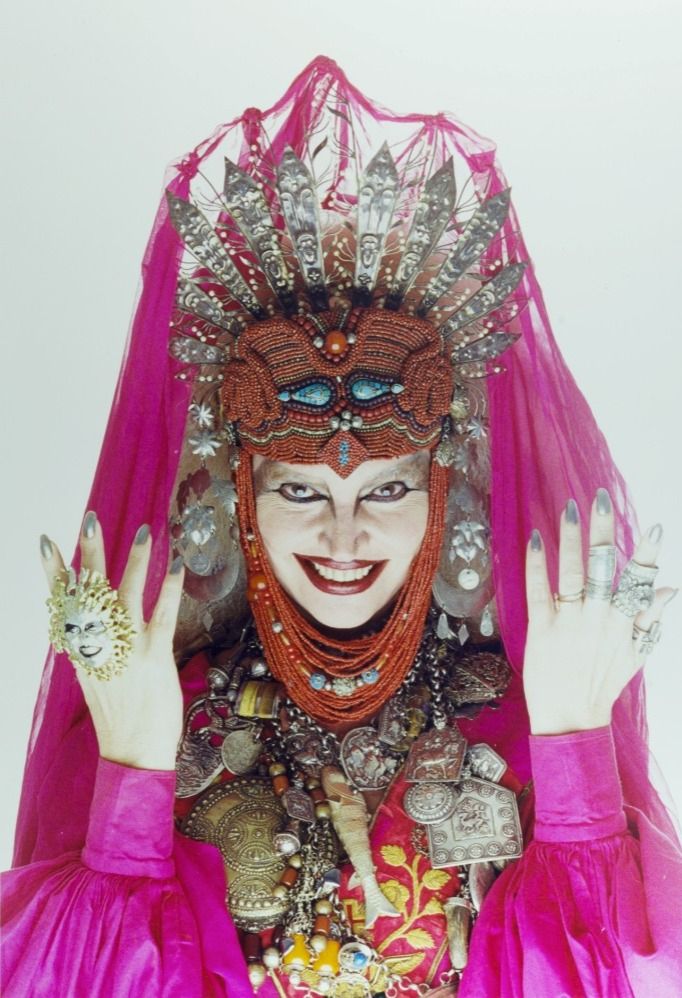 Elke Maravilha usando uma coroa elaborada com facas prateadas e miçangas vermelhas, um véu magenta, um colar com vários pendentes, vários anéis. Ela olha diabolicamente para a câmera.