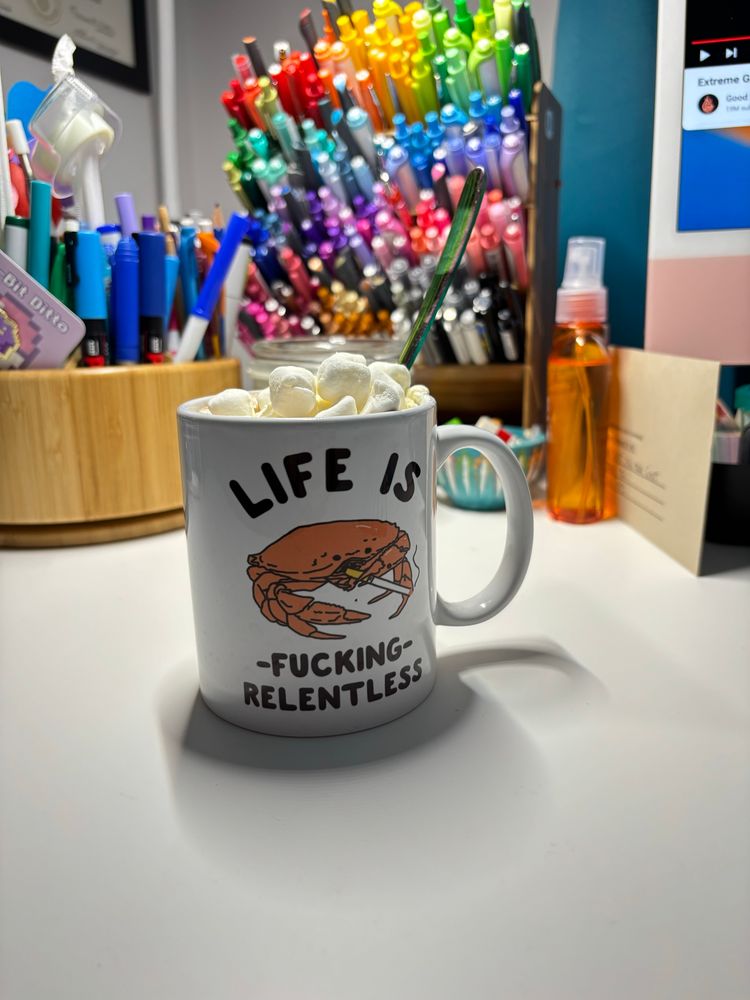 a hot drink with whipped cream and marshmallows in a mug. the mug has a crab smoking a cigarette and reads “life is fucking relentless” 