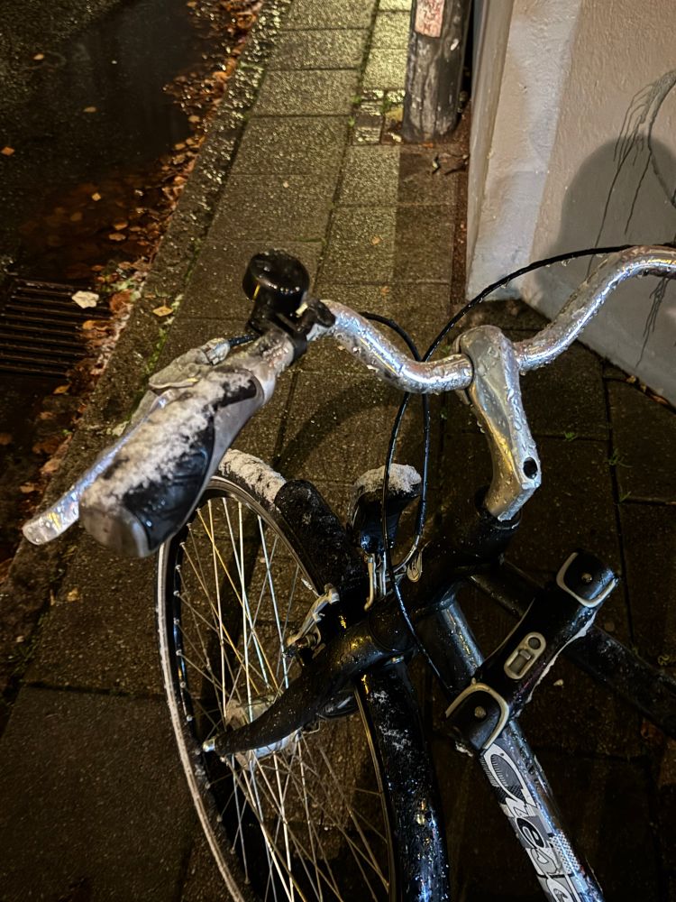 Ett cykelstyre, vänster handtag har lite snö på sig. Marken är våt men snöfri.