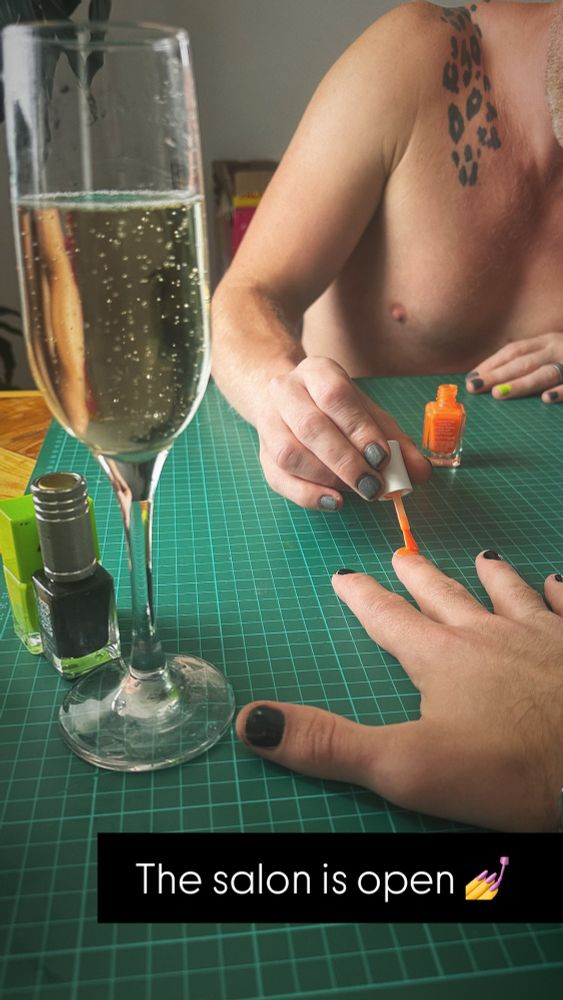 POV photo: my sitting at the kitchen table while my husband paints my nails black and orange. There’s a bubbly glass of prosecco to the side and a caption that says “THE SALON IS OPEN”