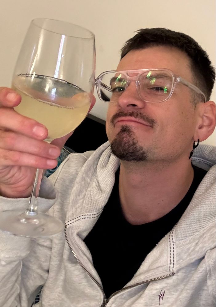 Selfie holiding a glass of white wine. I’m wearing a grey hoodie, black T-shirt and clear framed glasses. 