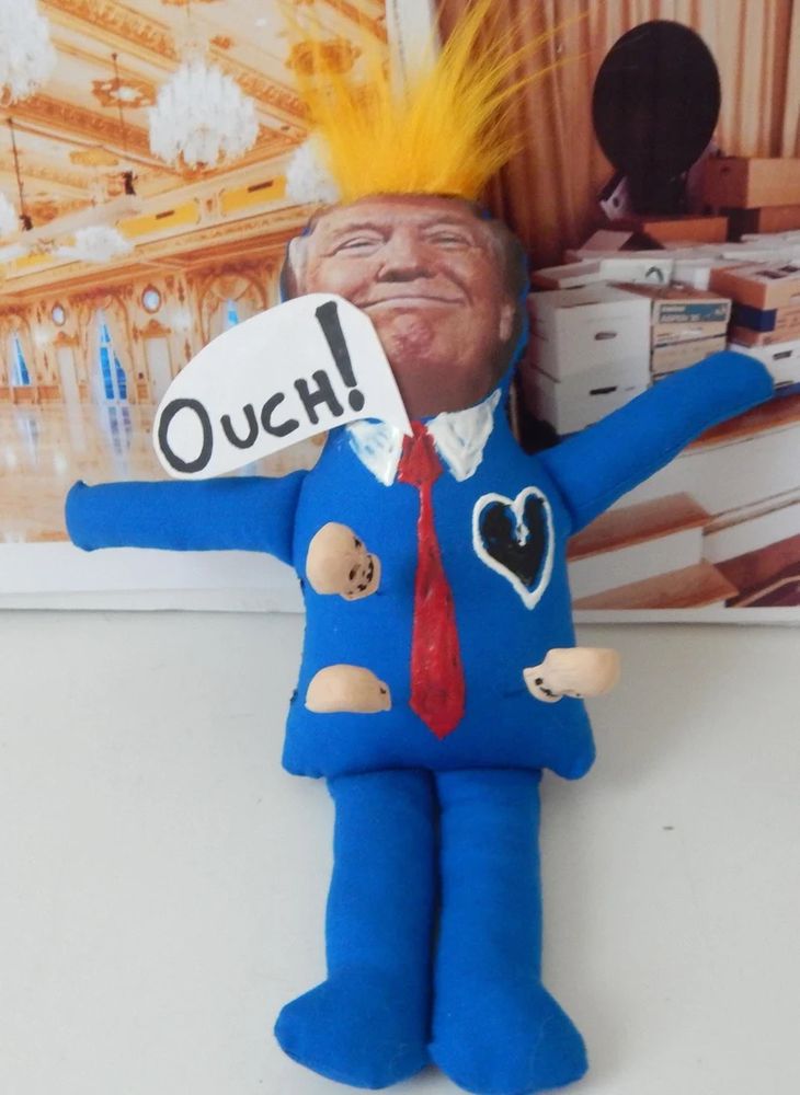 Voodoo doll with scull pins in a man with an orange face and blue suit with red tie. Backdrop is a gold adorned ballroom with chandeliers and a bunch of opened file boxes stacked up on a stage with a curtain backdrop. 
