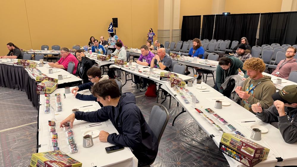 Wide shot of the room and attendees for a miniature painting workshop. Several people are sitting, spaced apart at tables, with a paint set in front of each of them. 