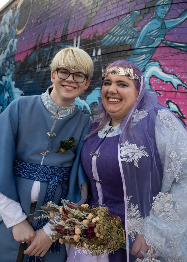 @werewulver.bsky.social and my dearly beloved but not yet on bsky friend Scribe. They are standing next to each other beaming in front of a purple-blue mural. Their handmade wedding outfits are vaguely elven: Nate's shirt is blue, with a high collar and a darker blue woven belt, and Scribe's dress is purple with a translucent veil with a mushroom motif over the top. They both look tremendously happy. 
