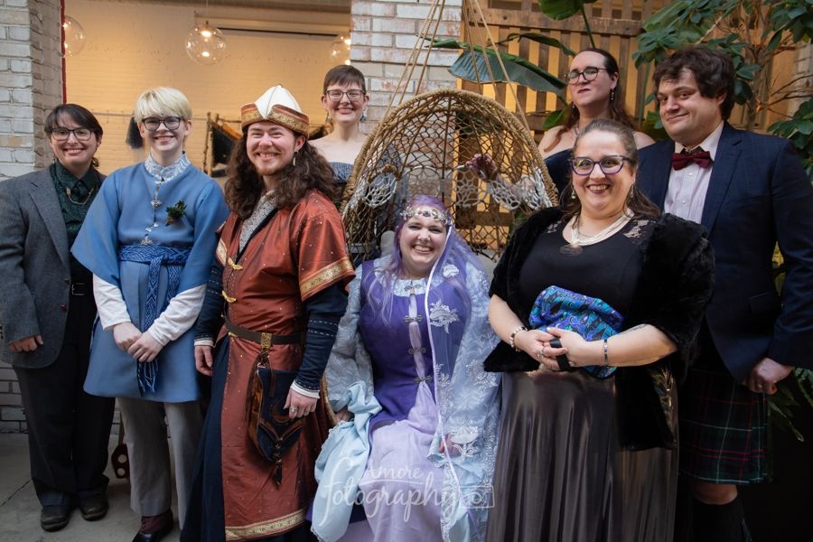 A cluster of people in various ridiculous and delightful outfits, all smiling widely, centered on Scribe in a hanging wicker chair and @werewulver.bsky.social standing nearby. @merovingians.bsky.social and I are two of the three people in the back row, looking and feeling like benevolent giants among a crowd of hobbits