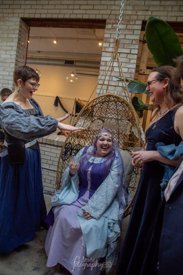 Scribe sitting enthroned in a hanging wicker chair with me and Em on either side, smiling widely. My arms are extended forward and it looks vaguely like I might be casting a spell; Em is laughing at me.