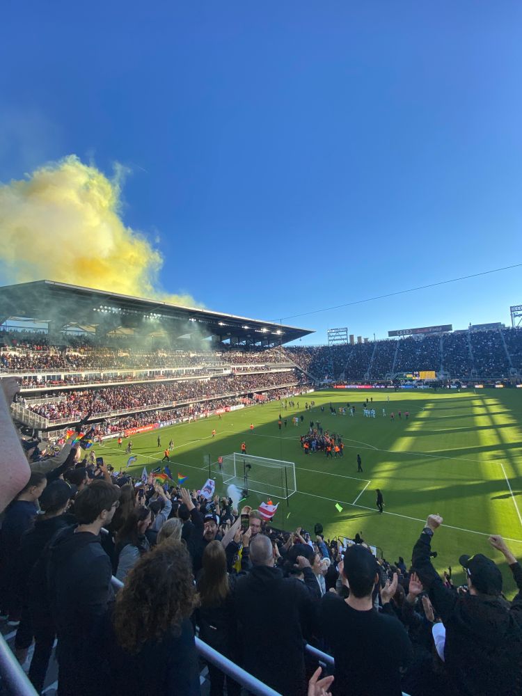 The Washington spirit team and staff celebrate on the field at Audi stadium, as a huge crowd cheers on the sidelines and yellow fireworks smoke fills the air. 