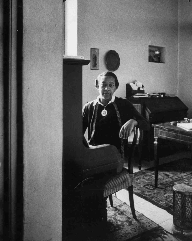 A black and white image of a woman sitting behind a piano in a furnished room as she looks at the camera.