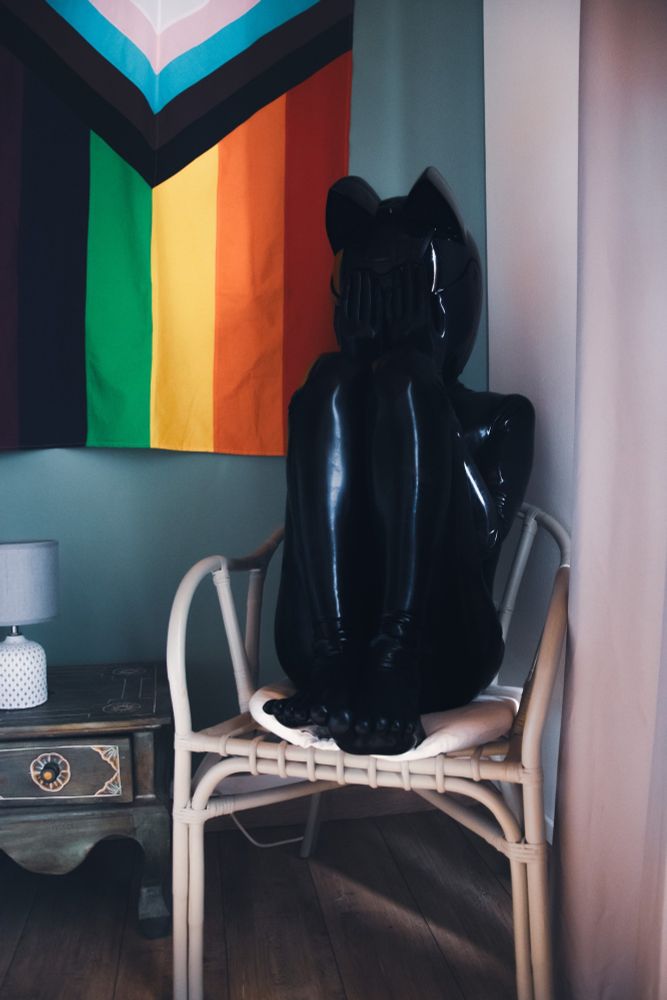 A person in full black catsuit latex, with toes socks and a helmet with car ears is curled up on a chair, putting its hands in front of his eyes, we can see behind it a pride flag