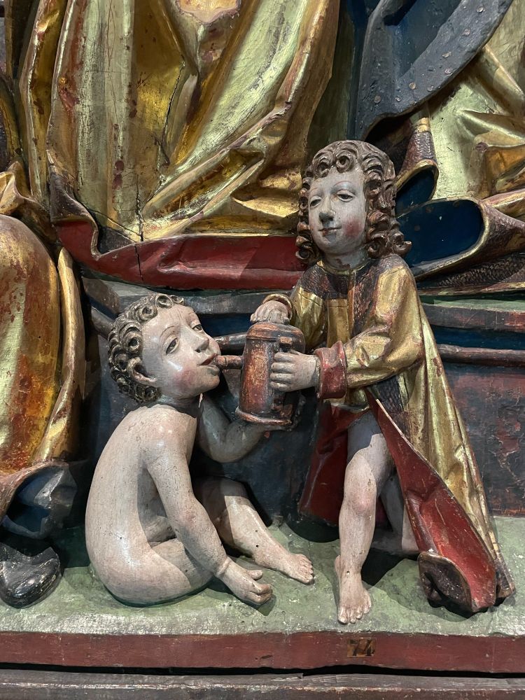 Statues, neither wearing pants. The person standing is holding a drink for the person sitting. 