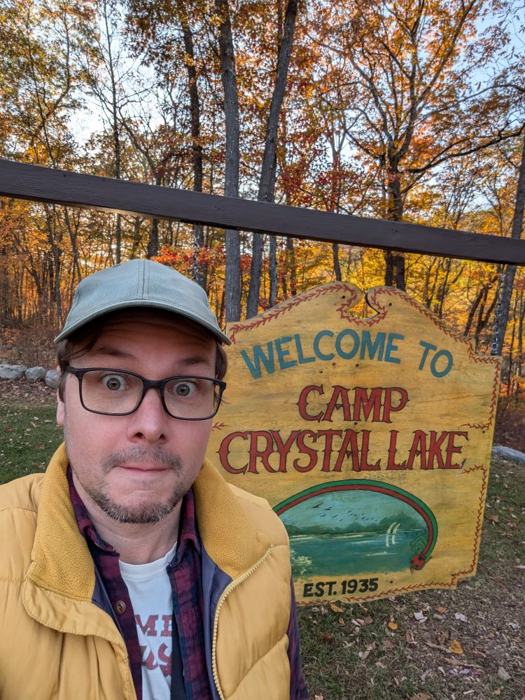 Me (middle-aged nerd, glasses, green baseball cap, yellow vest) standing in front of the Welcome to Camp Crystal Lake sign at Camp NoBeBoSco.