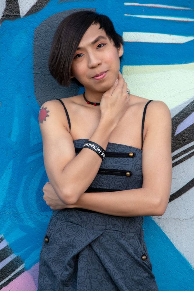 Kay facing the camera while smiling, head tilted with her left arm over her stomach and her right hand on her neck. In front of a primarily blue painted wall 