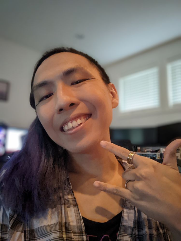 Kay smiling looking at the camera, left hand as ILY, head tilted with hair tied to her right 