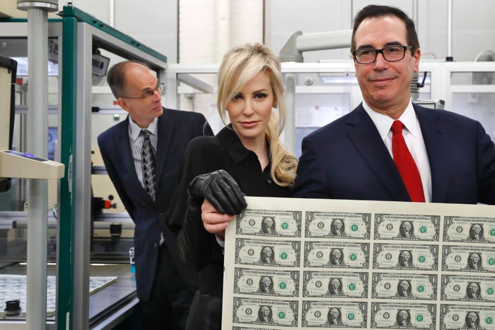a 2017 photo of Steven Mnuchin (right), actress Louise Linton (Mnuchin's wife), and bureau of engraving and printing director director Leonard Olijar (left). Linton and Mnuchin together hold up a freshly-printed sheet of dollar bills, him barehanded, her with full-length black leather gloves. She looks directly at the camera, head tilted down, bangs artfully half-covering one eye like a villainous Veronica Lake. Steve is also there, I guess