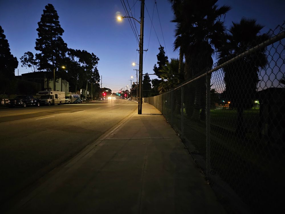 A chain link fence, a dark purple sky, pavement, the silhouettes of palm trees, a line of street lights, an empty road and sidewalk, all waiting for day to begin