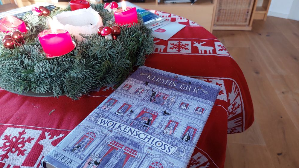 Auf einem Tisch mit einer rot - weißen Tischdecke mit Weihnachtsmotiv steht ein Adventskranz mit brennenden roten Kerzen. Davor liegt das Buch "Wolkenschloss".