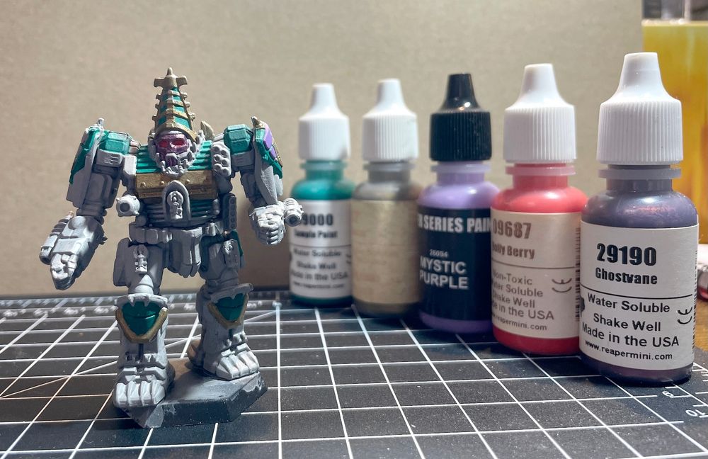A Battletech: Gothic miniature - an Atlas AS7-D, a giant, heavily armed mecha or robot, partially painted, standing next to a row of paint bottles from Reaper paints, labeled - Ghostvane, Holy Berry, Mystic Purple, Drillbit Metal, and a "Sample Paint" (Pthalo Green) - paints used to work on the Atlas. 