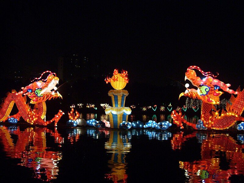 The Beijing Mid-Autumn Festival showing blown up lanterns of two dragons, a layered cake with a flaming ball at the top, all on top of water  in an urban area of the city