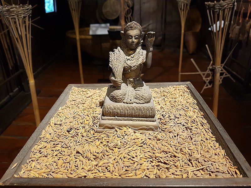 A statue of Phosop in Bangkok, showing a woman holding a sheaf of rice and holding her hand up; around the statue are large grains of dried rice.