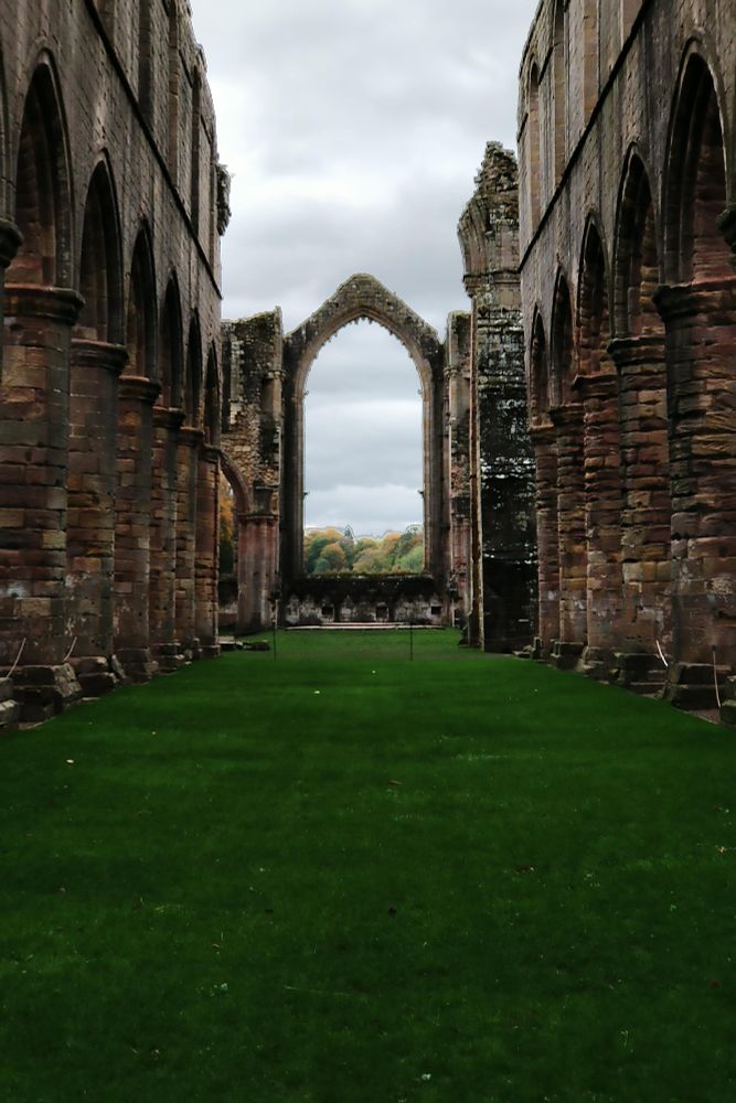 Fountains Abbey ruins of the nave with grass growing and no glass in the main window, but a view of the countryside instead, framed by the old stone.