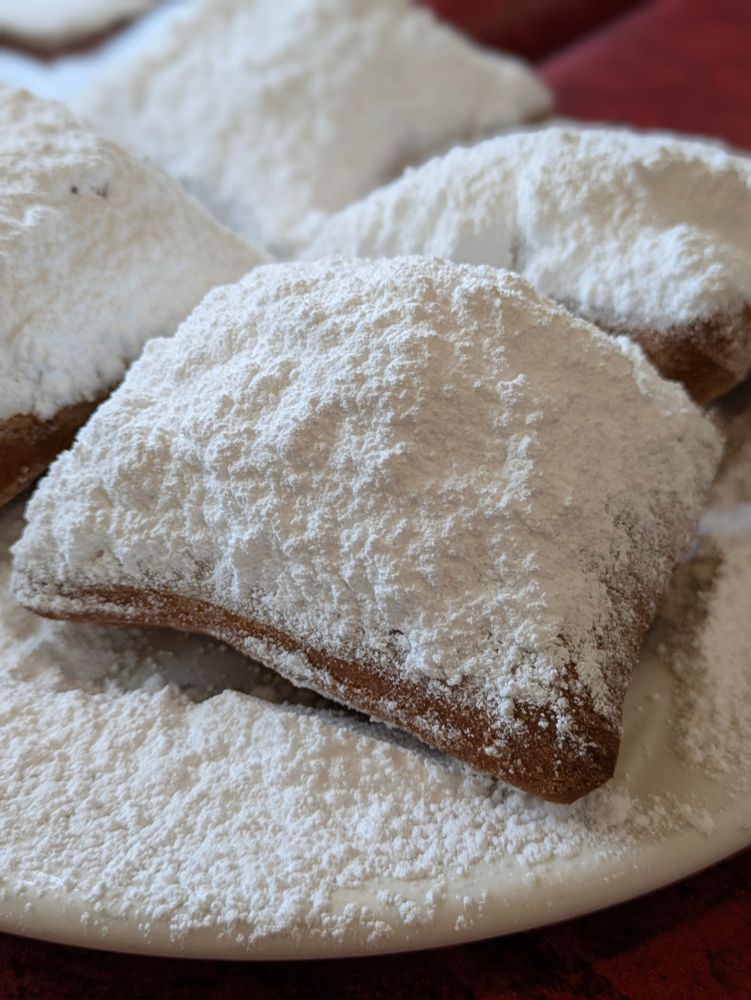 A white plate is covered in white powdered sugar. Beneath the sugar are four beignets, one in the foreground with the brown edges showing and three in the background. The plate is on a brown table top.