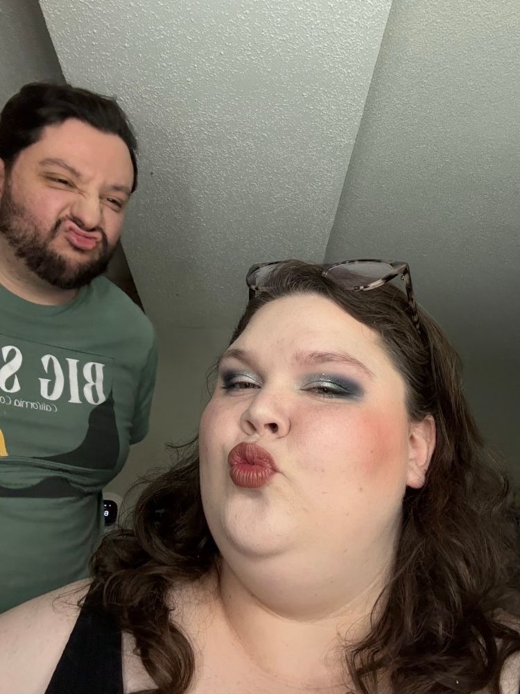 A selfie of a man to the back left of the image and a femme to the center right. He is wearing a green shirt with simple silhouettes of the ocean and rocks in black, with a yellow sun in the back. The text reads BIG SU- and is cut off. He has short black hair and short beard and mustache. The femme is wearing a black tank top, has brown curly hair, is wearing makeup, and glasses on top of her head like a head band. They are slightly turned to each other while still facing the camera and both are making kissy faces.