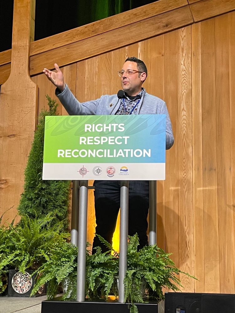 PICS Executive Director Ian Mauro speaks at a podium wearing a light blue blazer and dark blue, patterned shirt. His hand is raised in the air, elbow flexed, in a pointing gesture. A sign on the podium reads "RIGHTS. RESPECT. RECONCILIATION".
