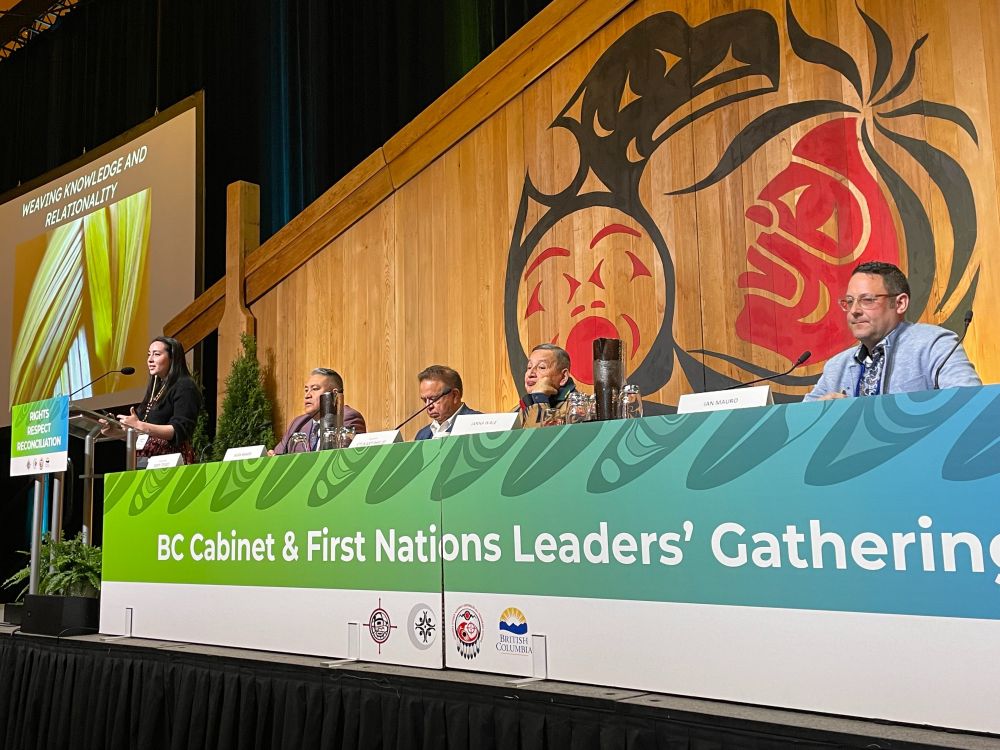 PICS Indigenous Research and Partnerships Lead Janna Wale speaks at a podium wearing a black top and a traditional ribbon skirt. A sign on the podium reads "RIGHTS. RESPECT. RECONCILIATION". To her right side, a screen reads "Weaving Knowledge and Relationality". To her left sits 
BC Assembly of First Nations Regional Chief Terry Teegee, Hugh Braker of the First Nations Summit, Grand Chief Stewart Phillip of the Union of B.C. Indian Chiefs, and PICS Executive Director Ian Mauro.