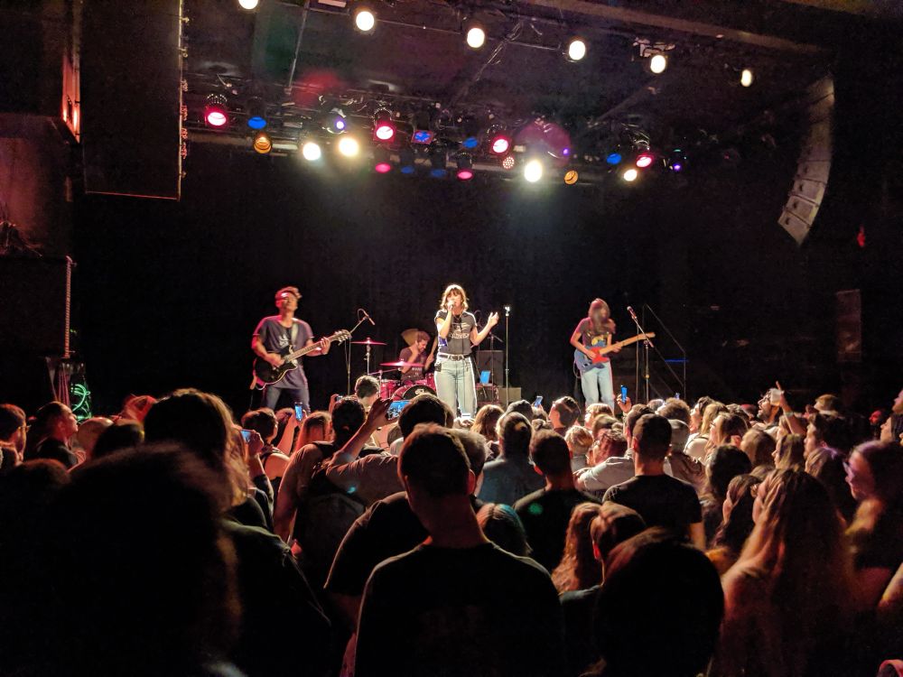 A packed audience watches Meg & Dia performing on stage at the Music Hall of Williamsburg in New York. The lead singer, Dia Frampton, stands in the center holding a microphone, gesturing with one hand while singing. To the left, a guitarist plays in a relaxed stance, and to the right, another band member, Meg Frampton, plays an electric guitar under the glow of stage lights. Bright white and colorful spotlights illuminate the band, contrasting with the dark background. The crowd in the foreground is dense, with many fans holding up their phones to capture the performance.
Credit @thecontraire.bsky.social. September 2019.