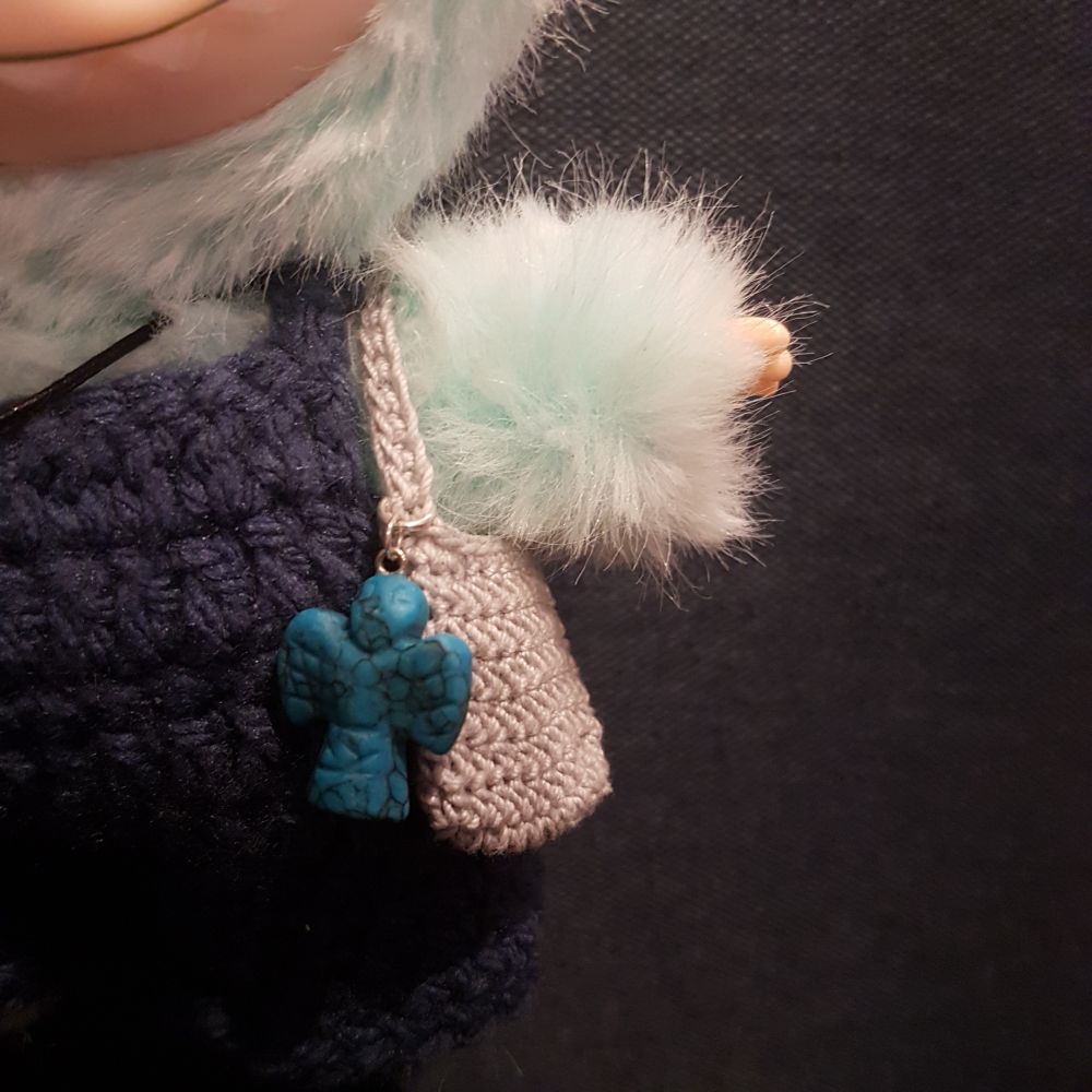 Close up of fluffy green toy's arm. He has blue dungarees, and a small grey crochet bag with a large blue angel hanging off it.
