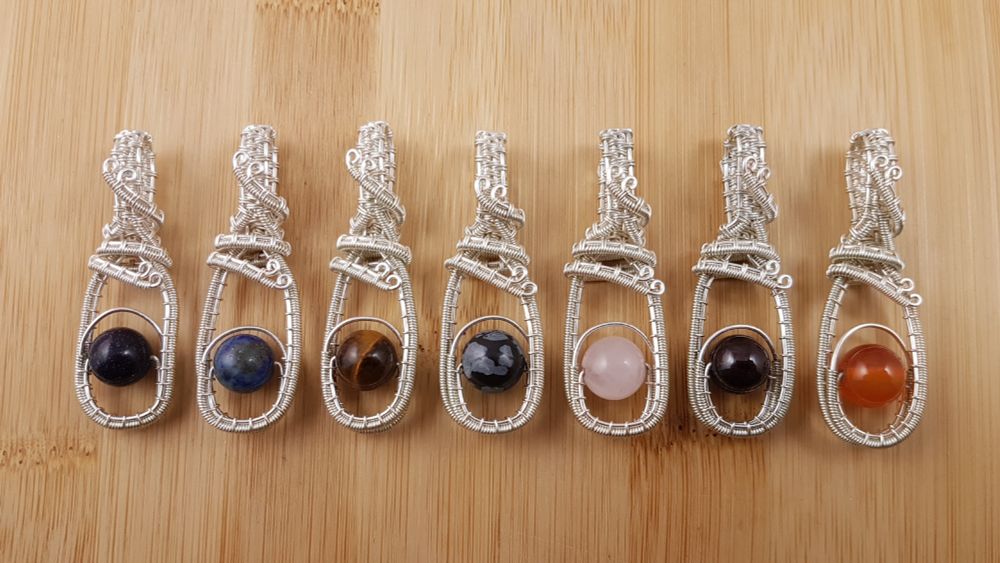 Row of 7 single gemstone bead and silver wire fidget pendants - sparkly blue, blue-green, brown, black and grey, pink, deep red, and orange.